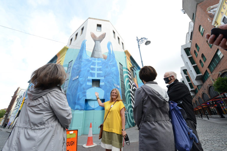 Group admiring vibrant mermaid mural on city building, colorful street art, bright day, cobblestone street.