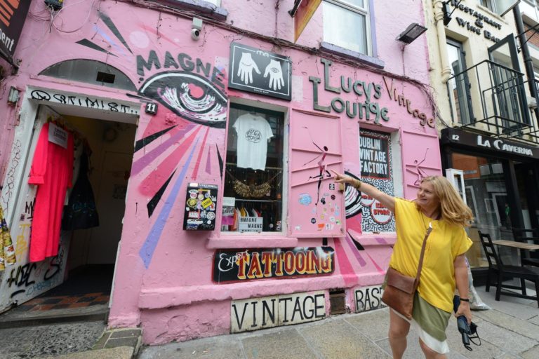 Woman pointing outside Lucy's Lounge, a vibrant pink vintage shop in Dublin with bold street art on the facade.