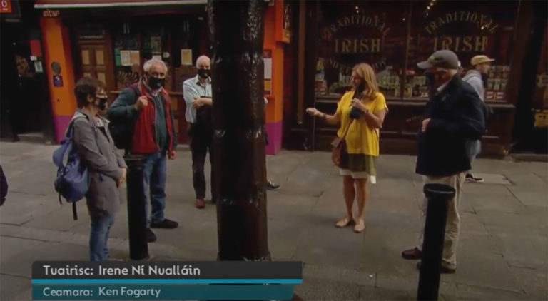 Group of people on a street tour outside a traditional Irish shop, wearing masks and listening to a guide.