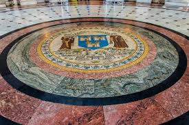 Mosaic emblem with intricate design on floor, featuring shield and figures, surrounded by patterned marble, in elegant setting.
