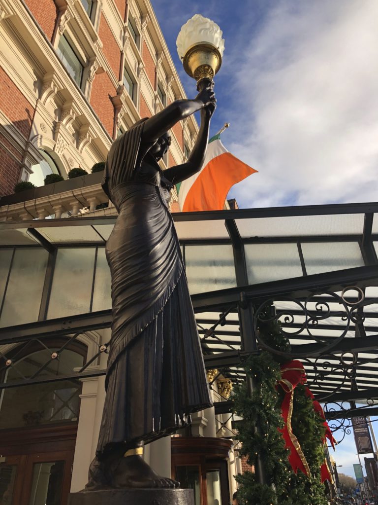 Bronze statue holding a lamp near a building with an Irish flag, Christmas decorations visible.