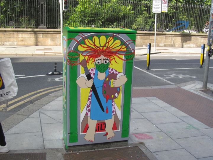 Colorful street art depicting a masked painter on a traffic utility box in an urban setting.