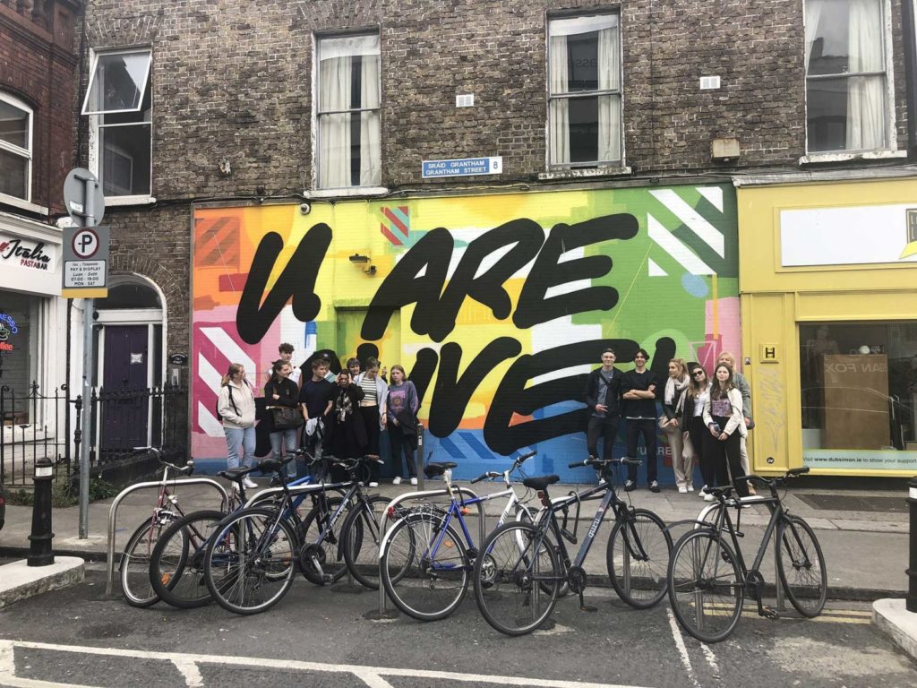 Group standing by colorful You Are Alive mural in urban street, with parked bikes in foreground.