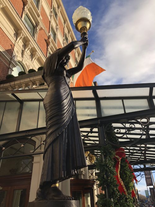 Bronze statue holding a lamp outside a historic brick building under a blue sky.