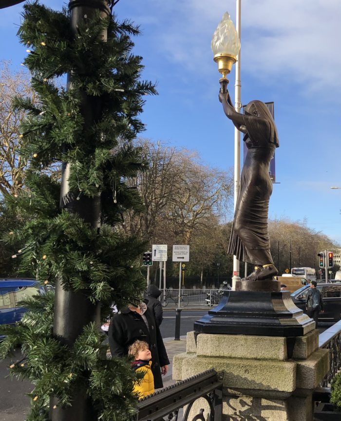 Bronze statue holding torch with festive wreath in foreground; child gazes up on a sunny street scene.