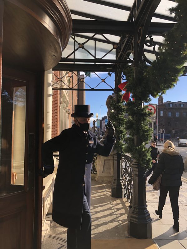 Doorman in black coat and top hat opening a door at a festively decorated entrance on a sunny day.