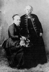 Two Victorian-era women in formal attire, one seated with a small dog, posing for a portrait.