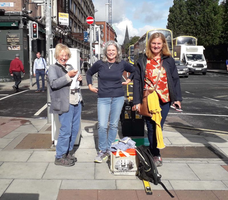 Three women smiling at a city street corner with shopping bags and a bus in the background.