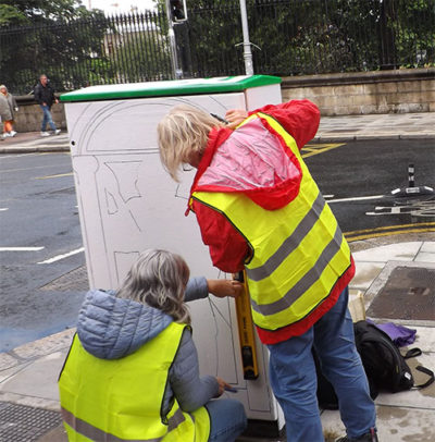 People wearing reflective vests painting a street utility box on a city sidewalk.