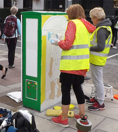 Artists in reflective vests painting a mural on a street utility box with vibrant colors.