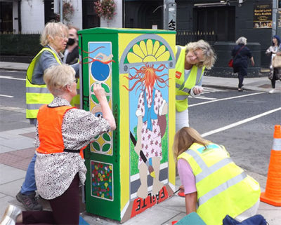 People painting a colorful mural on a street cabinet while wearing safety vests near an urban road.